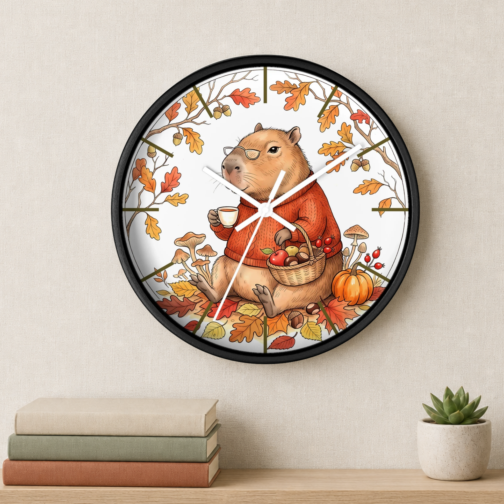 A round wall clock with a black frame hangs on a beige wall above a wooden shelf. The clock face features an autumn illustration of a capybara wearing glasses, holding a cup and basket of apples, surrounded by orange and yellow leaves, mushrooms, and a pumpkin.