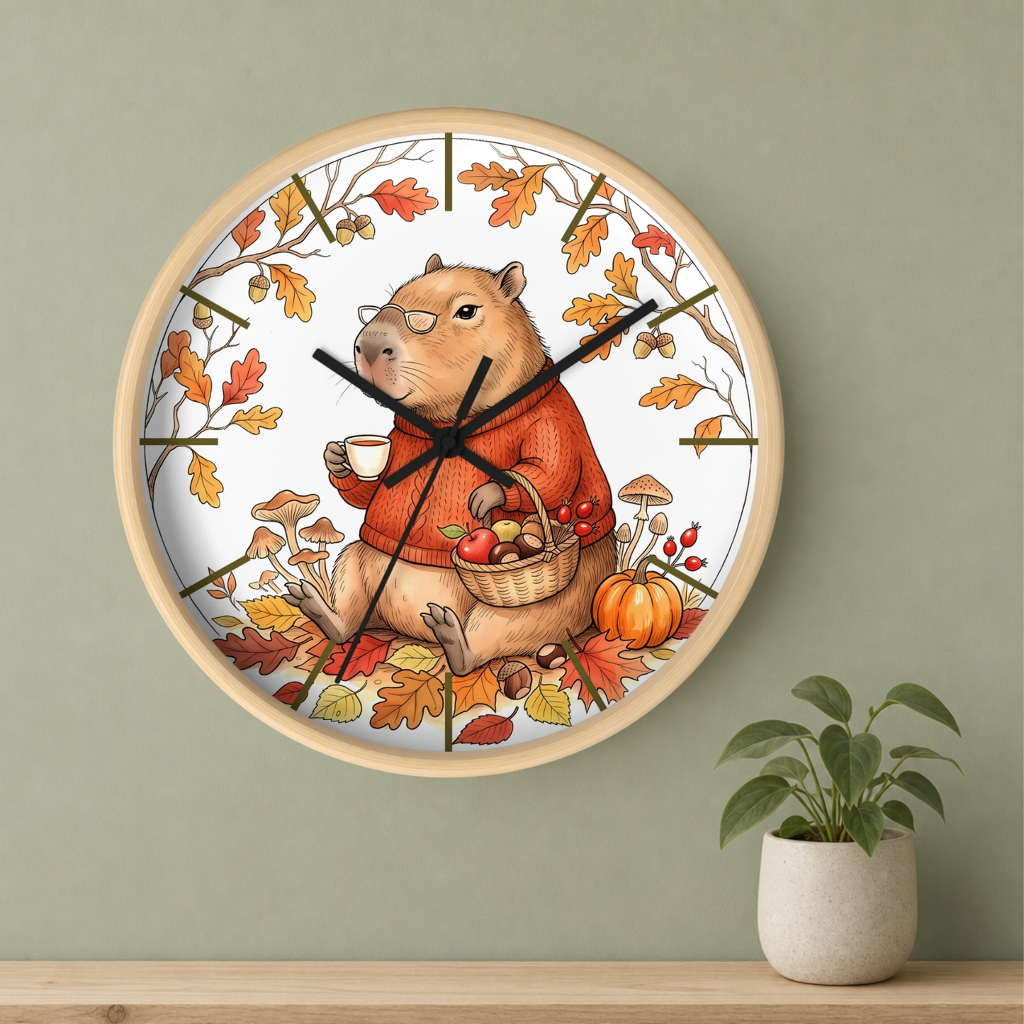 Round wall clock with a light wood frame, featuring an autumn-themed illustration of a capybara wearing an orange sweater and holding a basket of pumpkins and mushrooms. The clock is mounted on a sage green wall above a wooden shelf with a small potted plant.