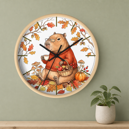 Round wall clock with a light wood frame, featuring an autumn-themed illustration of a capybara wearing an orange sweater and holding a basket of pumpkins and mushrooms. The clock is mounted on a sage green wall above a wooden shelf with a small potted plant.