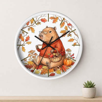 A round wall clock with a white frame and a whimsical autumn illustration of a capybara in an orange sweater, sitting among fall leaves, pumpkins, and mushrooms. The clock is mounted on a light beige wall above a wooden surface with a small potted succulent.