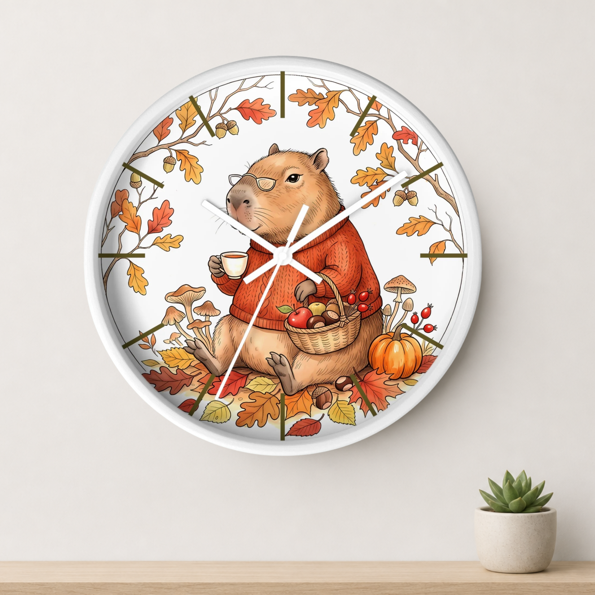 A round wall clock with a white frame and a whimsical illustration of a capybara dressed in an orange sweater, surrounded by autumn leaves, pumpkins, and mushrooms. The clock is mounted on a light beige wall above a wooden shelf with a small potted succulent.