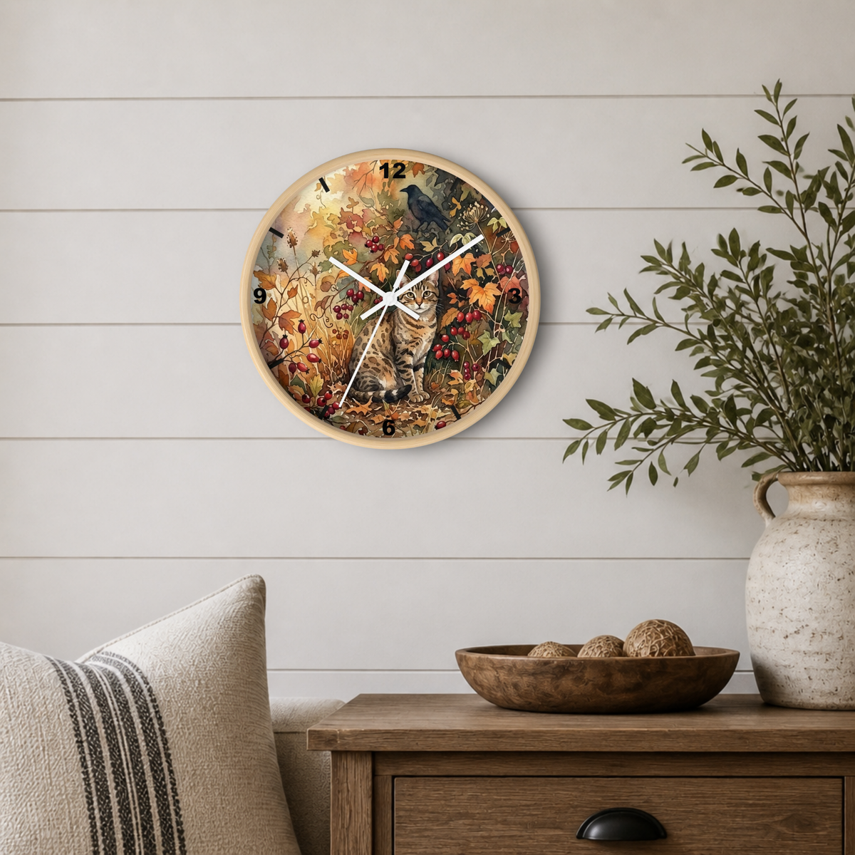 A round wall clock with a light wood frame hangs on a white shiplap wall above a wooden side table. The clock face features a detailed autumn scene with a cat, bird, and fall foliage.