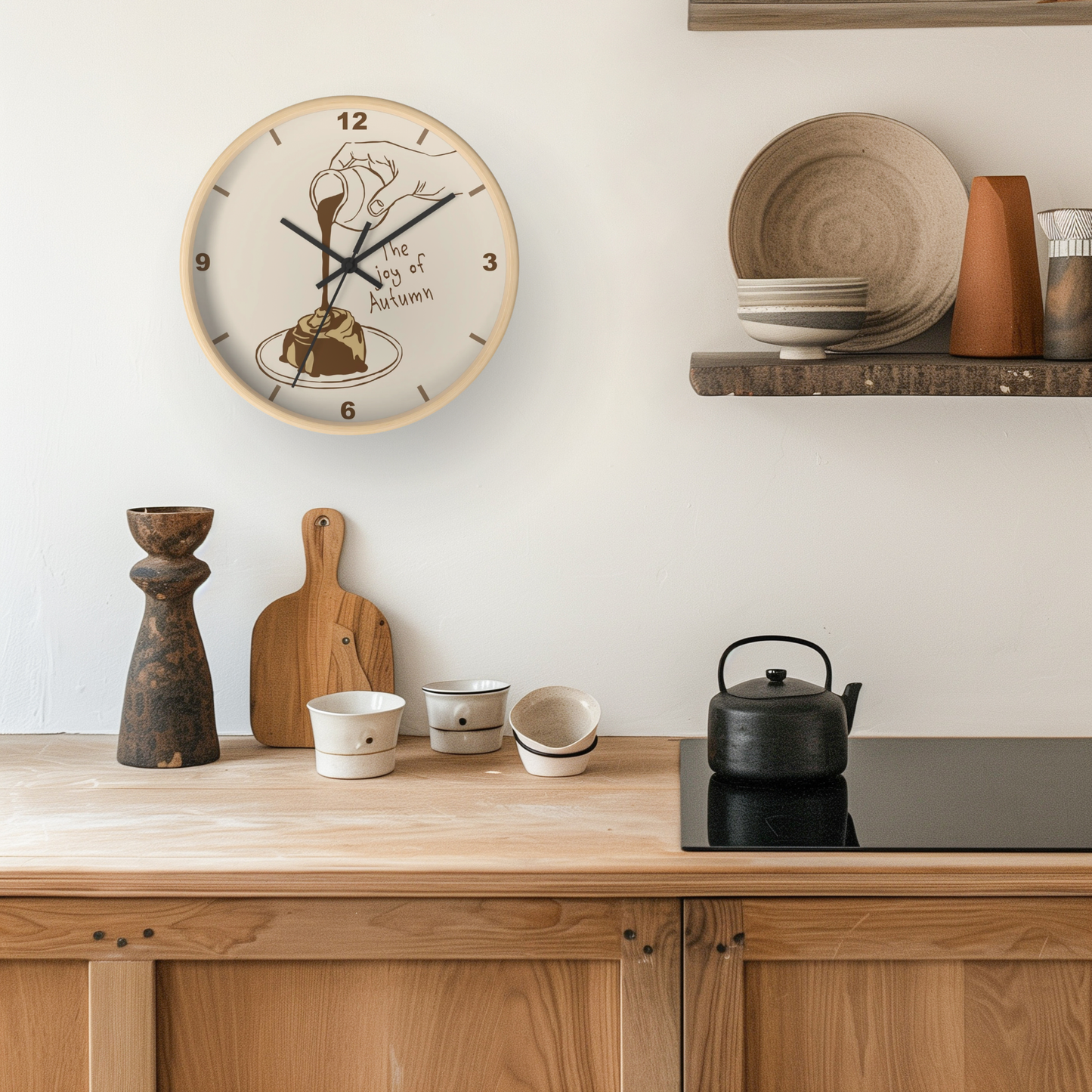 A round wall clock with a cream face and light wood rim hangs on a white wall above a wooden kitchen counter. The clock features a stylized illustration of a person holding a cup and saucer with the text 'The Day of Autumn'.