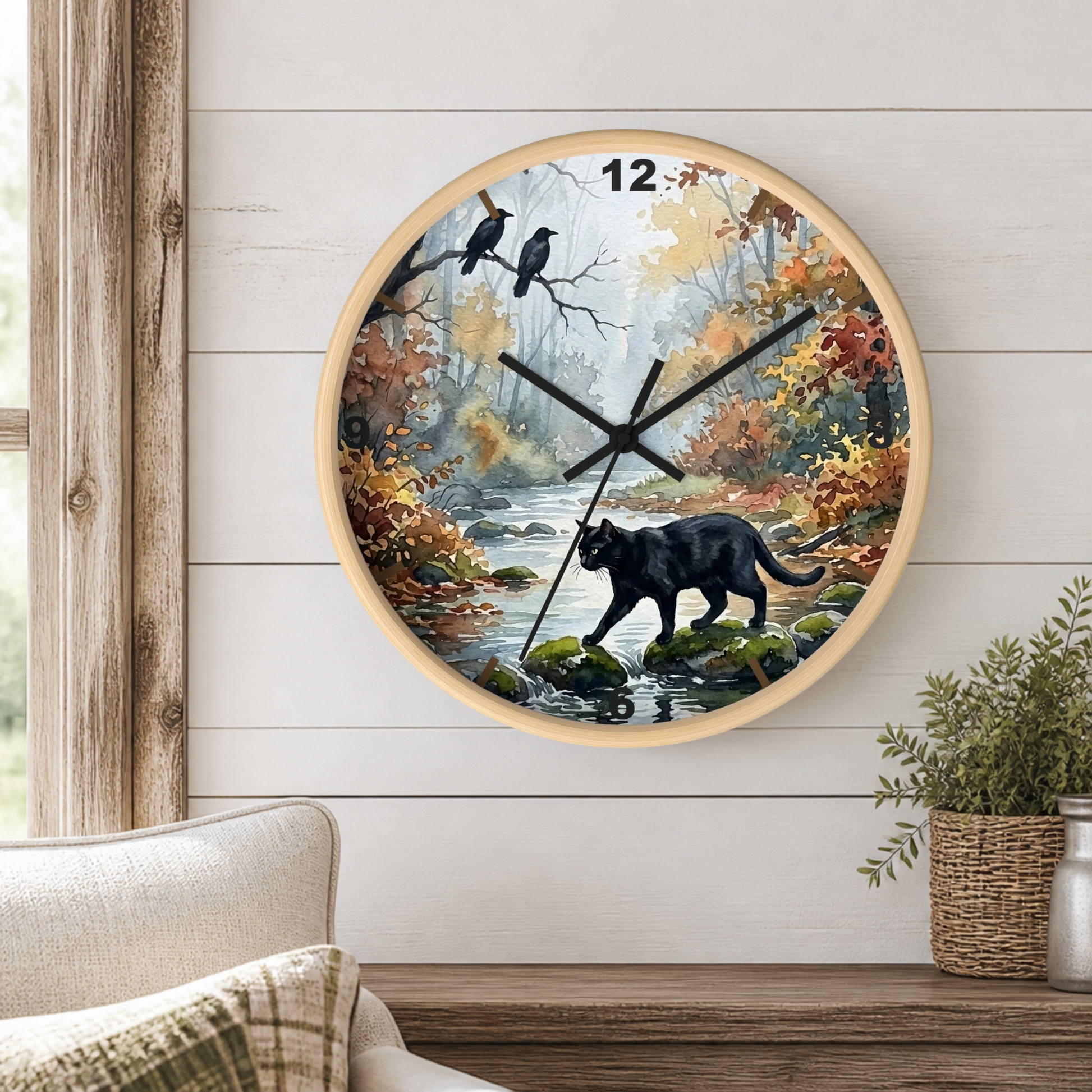 A round wall clock with a light wood frame hangs on a white shiplap wall above a wooden console table. The clock face features a scenic autumn forest illustration with a black cat walking by a stream, two crows on a branch, and the number 12 at the top.