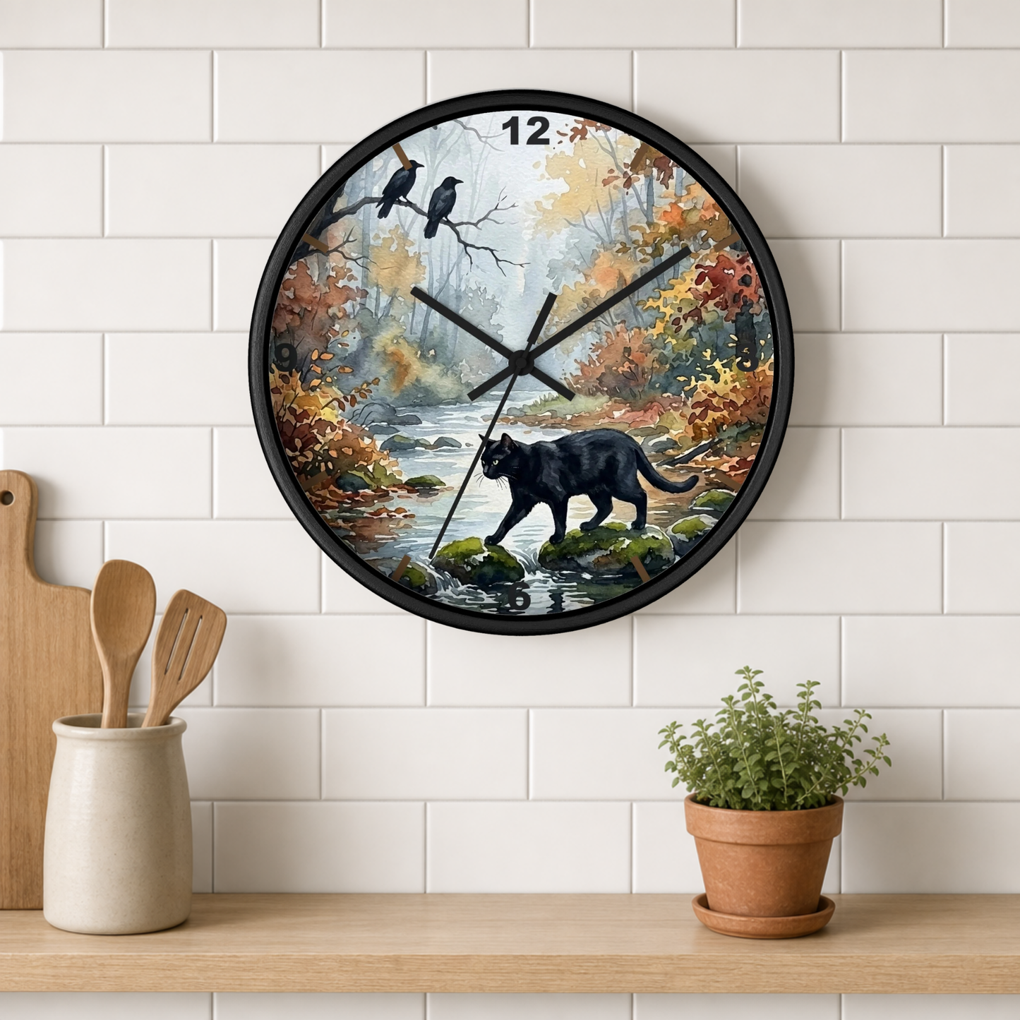 A round wall clock with a black frame hangs on a white subway tile wall above a wooden shelf. The clock face features a scenic autumn forest illustration with a black cat walking by a stream, two birds on a branch, and colorful foliage.