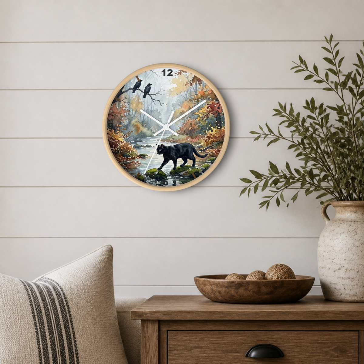A round wall clock with a nature illustration of a black bear by a river, mounted on a white shiplap wall above a wooden side table with a plant and decorative bowl.