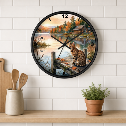 A round wall clock with a black frame hangs on a white subway tile wall above a wooden shelf. The clock face features a scenic autumn landscape with a lake, cabin, dock, and a spotted cat sitting on the dock.