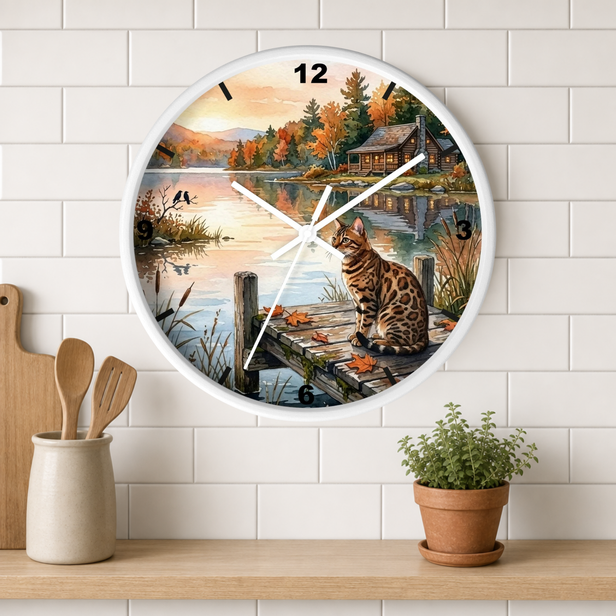 A round wall clock with a white frame hangs on a white subway-tile wall above a wooden shelf. The clock face features a scenic autumn lakeside print with a cat on a dock, a cabin, and fall foliage.