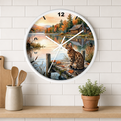 A round wall clock with a white frame hangs on a white subway-tile wall above a wooden shelf. The clock face features a scenic autumn lakeside print with a cat on a dock, a cabin, and fall foliage.