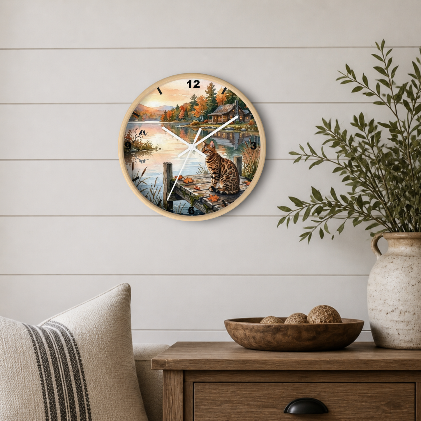 Round wall clock with a scenic autumn landscape print featuring a cat by a lake, mounted on a white shiplap wall above a wooden side table with a plant and decorative bowl.