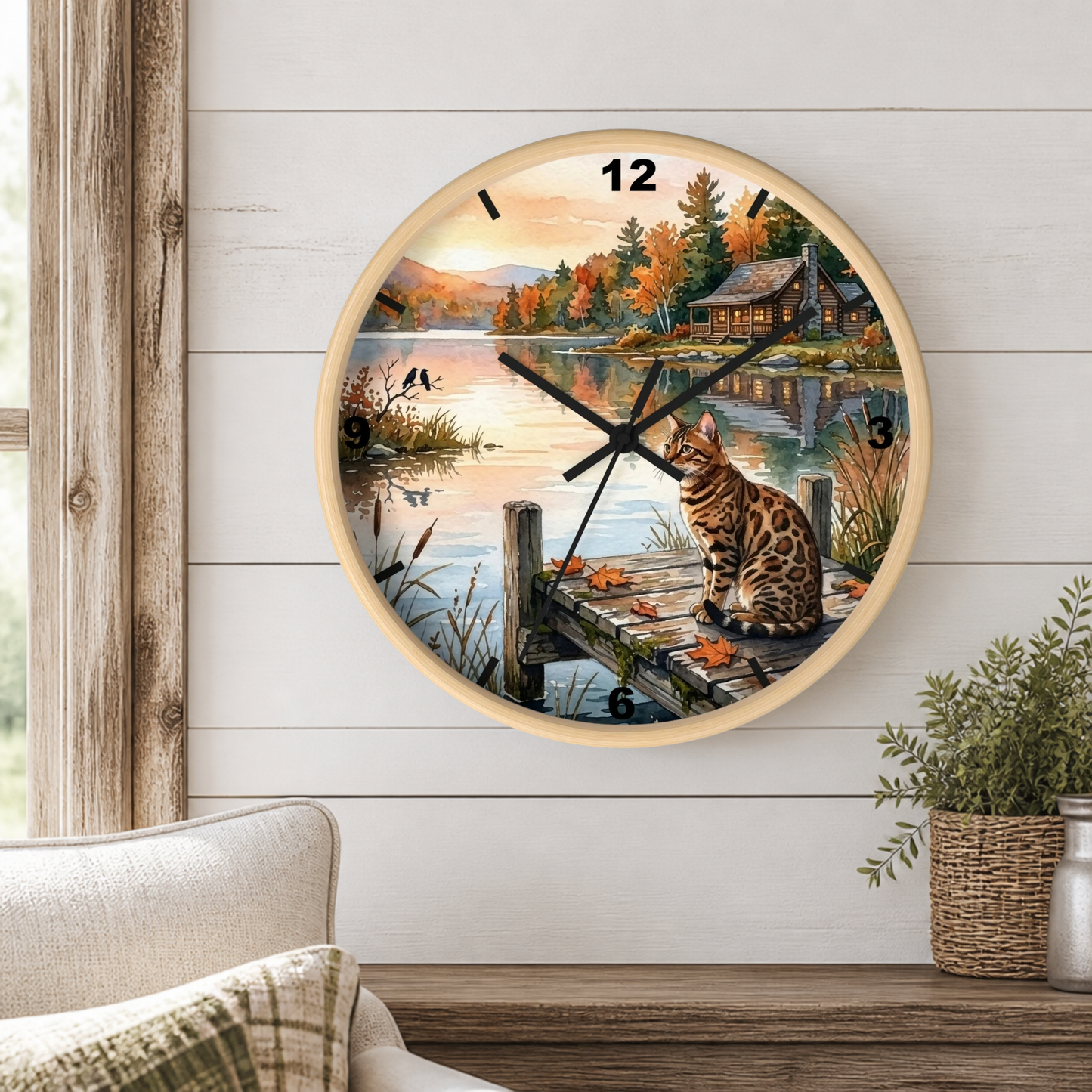 A round wall clock with a light wood frame hangs on a white shiplap wall above a wooden table. The clock face features a scenic illustration of a cat sitting on a dock by a lake, with autumn trees, a cabin, and mountains in the background.
