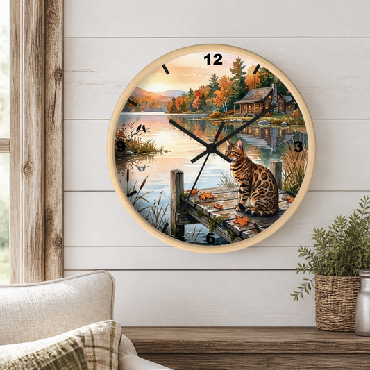 A round wall clock with a light wood frame hangs on a white shiplap wall above a wooden table. The clock face features a scenic illustration of a cat sitting on a dock by a lake, with autumn trees, a cabin, and mountains in the background.