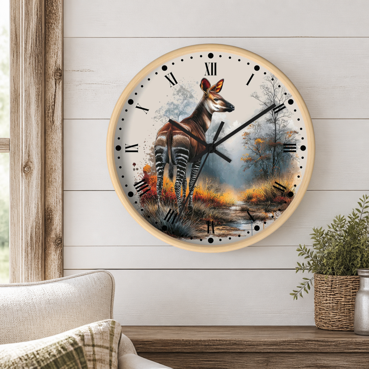 A round wall clock with a light wood frame hangs on a white shiplap wall above a wooden table. The clock face features a detailed illustration of a zebra deer standing in a misty forest landscape with autumn foliage.