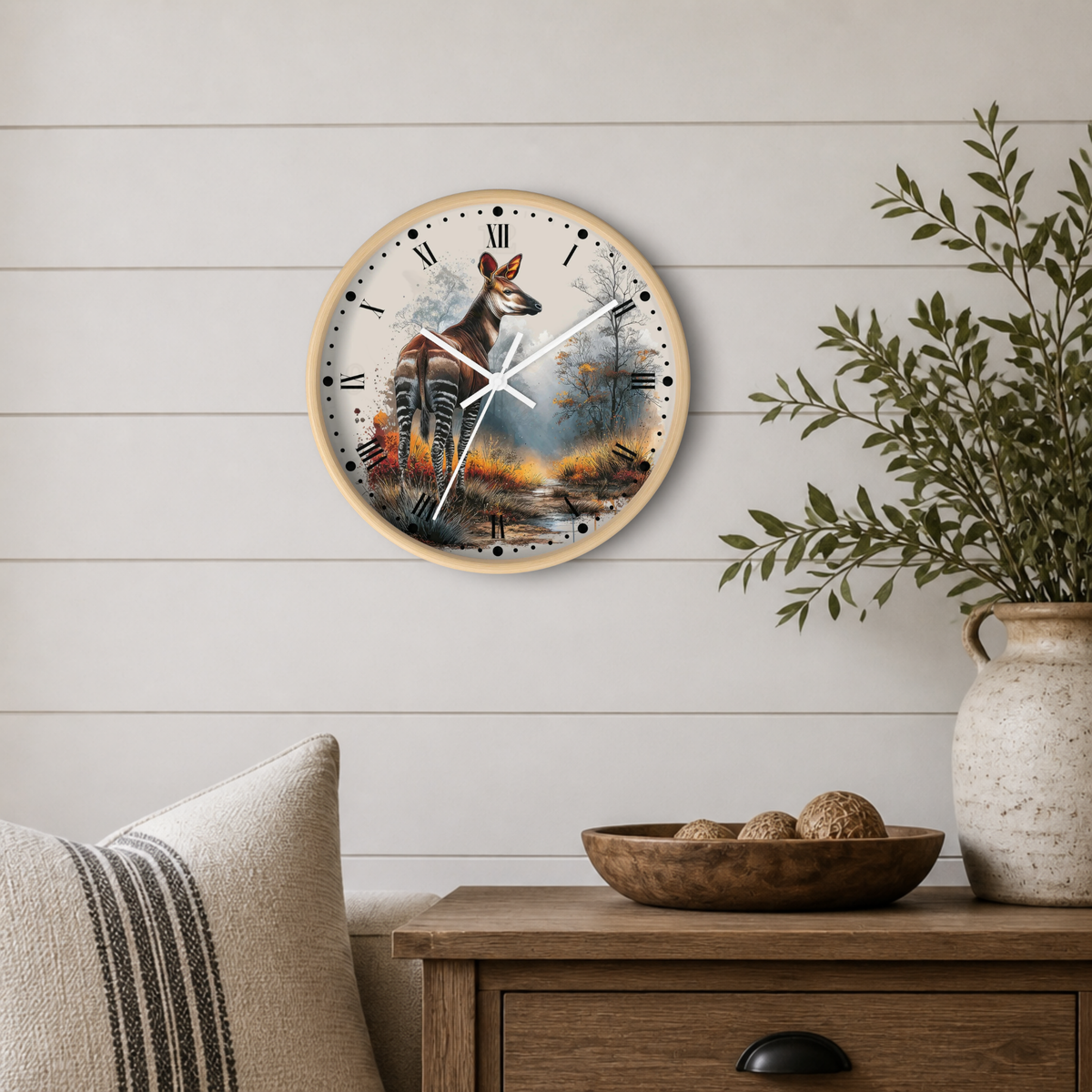 A round wall clock with a white frame and a zebra deer illustration on the face, mounted on a light wood shiplap wall above a wooden side table with a bowl and a vase of greenery.