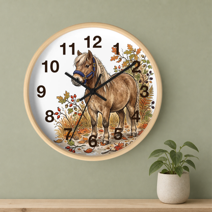 Round wall clock with a light wood frame and a white face featuring an illustrated brown horse surrounded by autumn leaves. The clock is mounted on a sage green wall above a wooden shelf with a small potted plant.