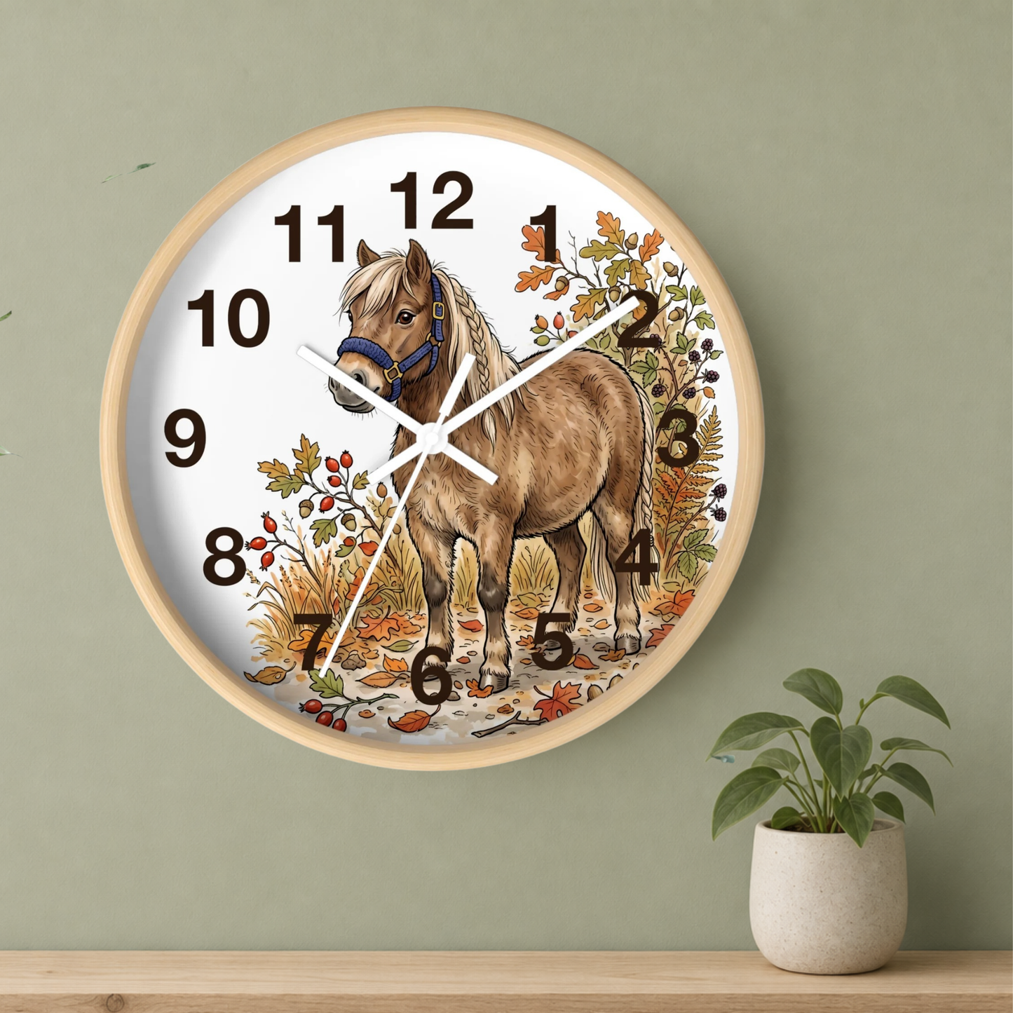 A round wall clock with a light wood frame and a white face, featuring an illustration of a brown horse surrounded by autumn leaves. The clock is mounted on a sage green wall above a wooden shelf with a small potted plant.