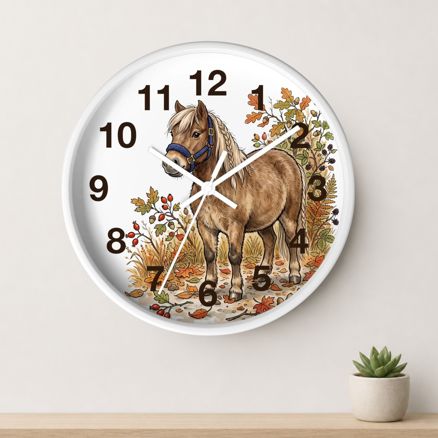 Round wall clock with a white face and rim, featuring an illustration of a brown horse surrounded by autumn leaves and berries. The clock is mounted on a light beige wall above a wooden shelf with a small potted succulent.