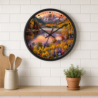 A round wall clock with a black frame and a scenic autumn landscape print, mounted on a white subway tile wall above a wooden shelf with kitchen utensils and a potted plant.