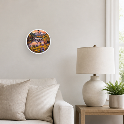 A round wall clock with a colorful landscape print is mounted on a light beige wall in a styled living room setting next to a white table lamp and a small potted plant.