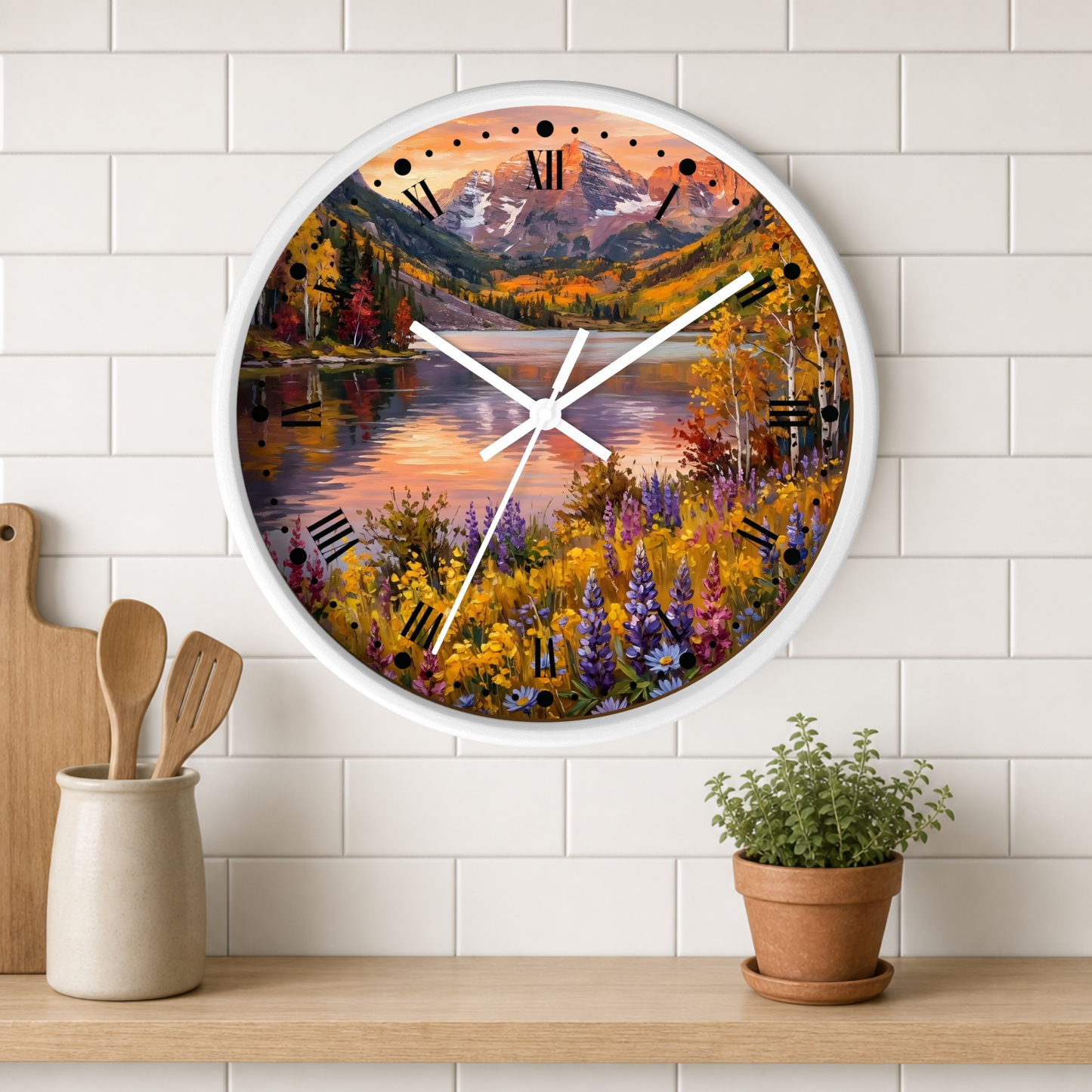 A round wall clock with a white frame hangs on a white subway-tile kitchen wall above a wooden shelf. The clock face features a colorful autumn landscape painting with mountains, a lake, and wildflowers.