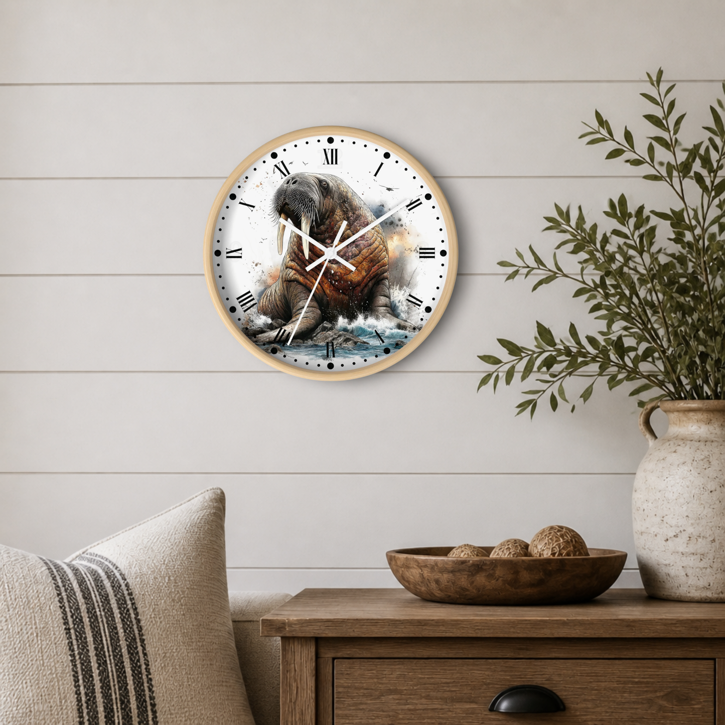 Round wall clock with a light wood frame featuring a detailed illustration of a walrus on the front, styled on a white shiplap wall above a wooden side table with a plant and decorative bowl.