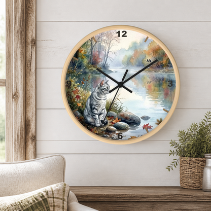 A round wall clock with a light wood frame hangs on a white shiplap wall above a wooden mantel. The clock face features a painted autumn landscape with a gray tabby cat sitting by a river, colorful fall foliage, and a misty forest scene.