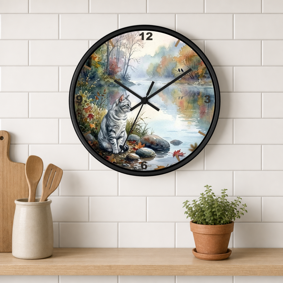 A round wall clock with a black frame and a scenic autumn landscape print featuring a gray tabby cat sitting by a river, mounted on a white subway tile wall above a wooden shelf with kitchen utensils and a potted plant.