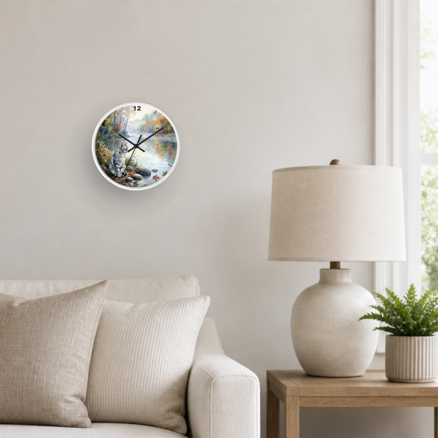 A round wall clock with a colorful nature scene print hangs on a light beige wall above a sofa. A white ceramic table lamp and a small potted plant sit on a wooden side table next to a window.