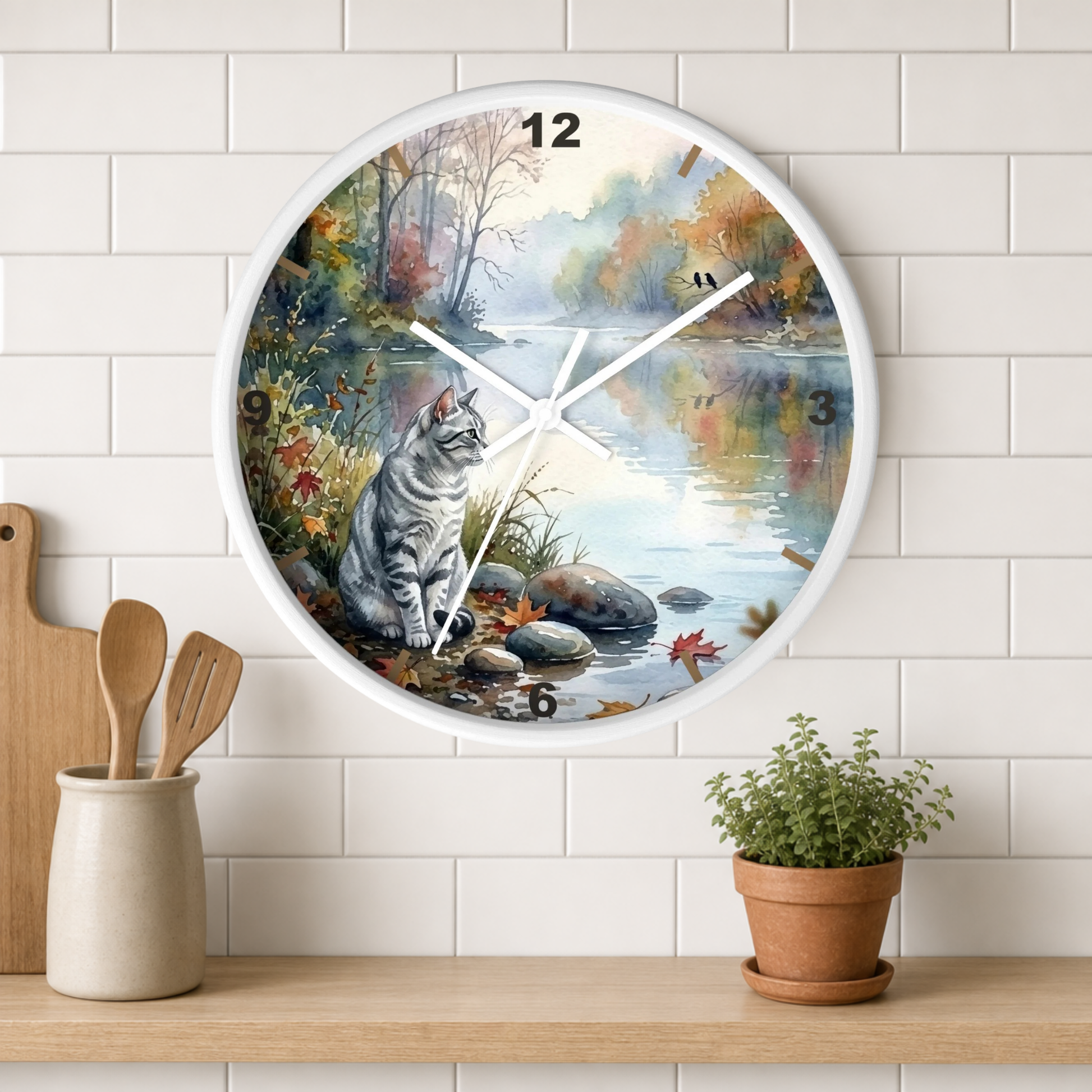A round wall clock with a white frame hangs on a white subway-tile wall above a wooden shelf. The clock face features a painted scene of a gray tabby cat sitting by a river with autumn trees and water reflections.
