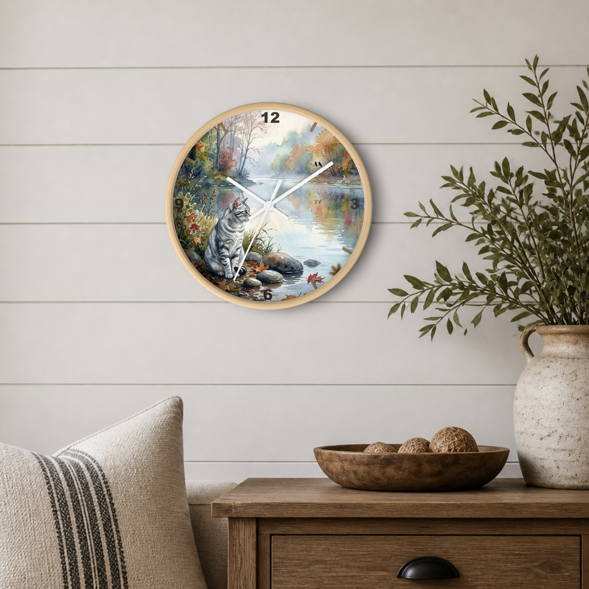 A round wall clock with a light wood frame hangs on a white shiplap wall above a wooden side table. The clock face features a painted autumn landscape with a gray tabby cat sitting by the water, colorful foliage, and a blue sky.