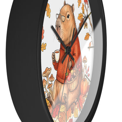 A round wall clock with a black frame and a whimsical illustration of a capybara wearing glasses and an orange sweater, holding a mug and a basket of autumn produce, surrounded by leaves and mushrooms.