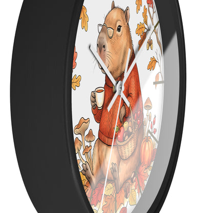 A round wall clock with a black frame and a whimsical illustration of a capybara wearing glasses and an orange sweater, holding a mug and a basket of autumn produce, surrounded by leaves and mushrooms.