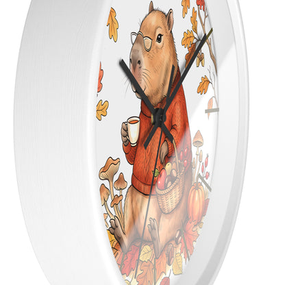 A round white wall clock featuring a whimsical illustration of a capybara wearing glasses and an orange sweater, holding a mug and a basket of autumn produce among falling leaves.