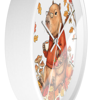 A round white wall clock featuring a whimsical illustration of a capybara wearing round glasses, a rust-orange sweater, and holding a mug while surrounded by autumn leaves, mushrooms, and pumpkins.
