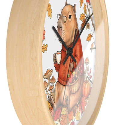 Round wall clock with a light natural wood frame and a whimsical illustration of a capybara wearing glasses and an orange sweater, holding a mug and a basket of autumn produce, on a white background.