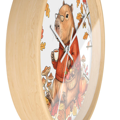 Round wall clock with a light natural wood frame and a whimsical illustration of a capybara wearing glasses and an orange sweater, holding a mug and a basket of autumn produce, on a white background.