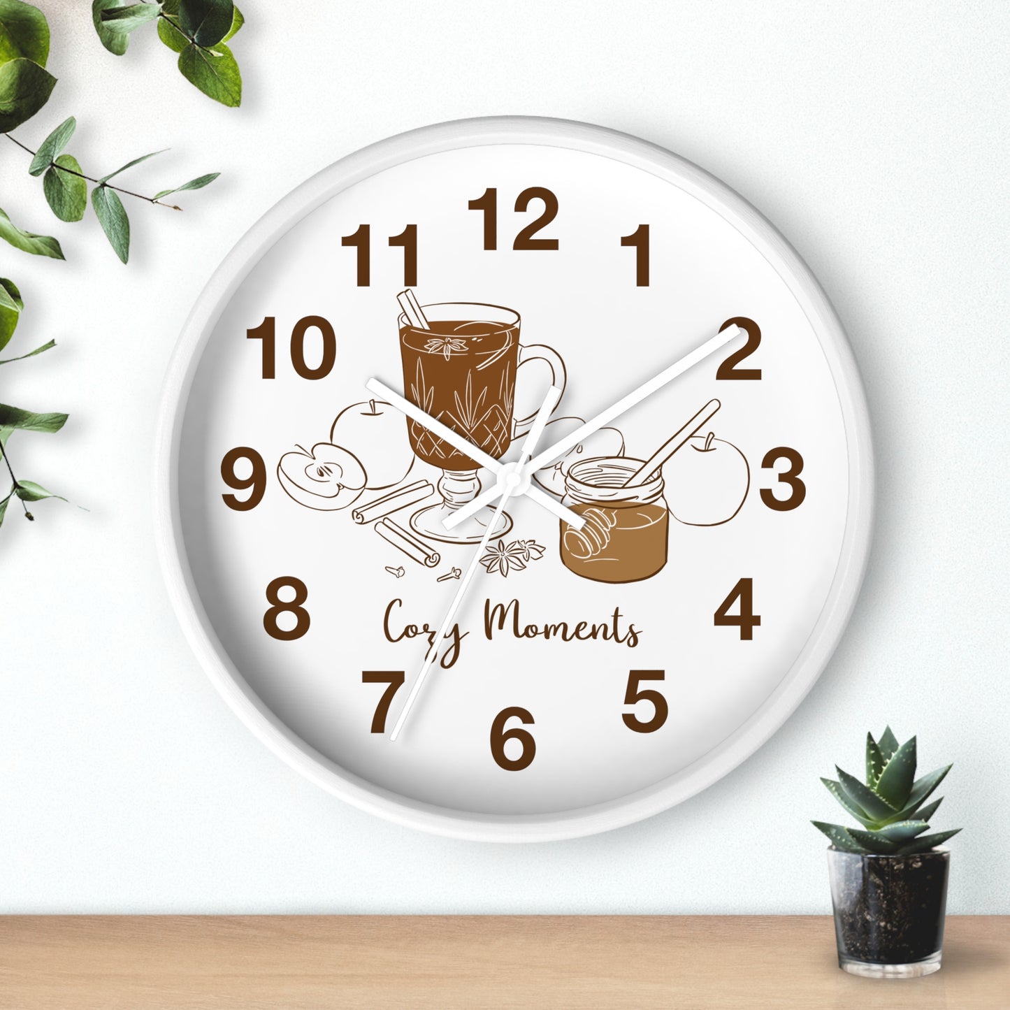 A round white wall clock with a warm-toned illustration of a mug, cinnamon sticks, and jam on the face, with the words 'Cozy Moments' and brown numerals. The clock is shown hanging on a white wall above a light wood surface, with decorative green leaves and a small potted succulent nearby.