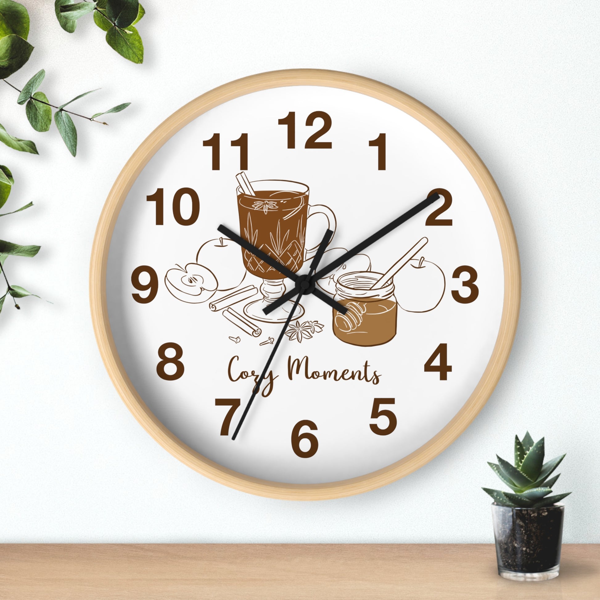 A round wall clock with a light wood frame and a white face, featuring brown numerals and illustrations of a coffee cup, cookies, and the words 'Cozy Moments.' The clock is shown hanging on a white wall above a wooden surface, with small green plants in the corners.