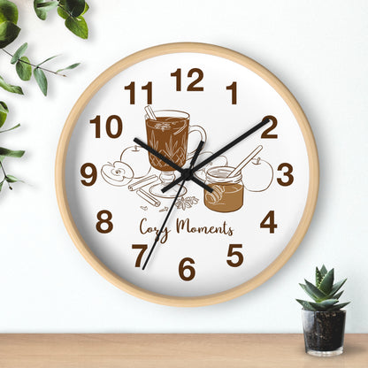 A round wall clock with a light wood frame and a white face, featuring brown numerals and illustrations of a coffee cup, cookies, and the words 'Cozy Moments.' The clock is shown hanging on a white wall above a wooden surface, with small green plants in the corners.