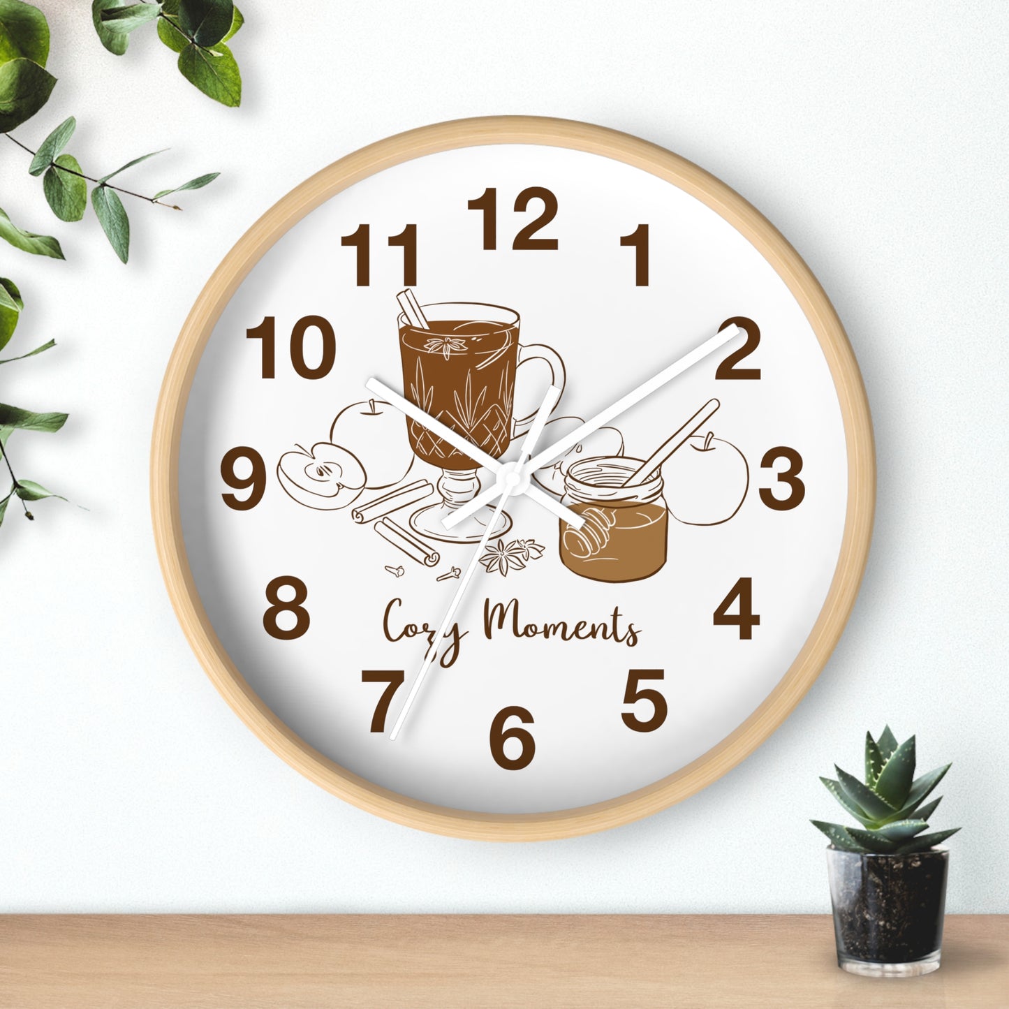 A round wall clock with a light wood frame and a white face, featuring brown numerals and illustrations of a mug, cinnamon sticks, and a jar with the words 'Cozy Moments' on the clock face. The clock is mounted on a white wall above a wooden surface, with decorative green leaves and a small potted succulent nearby.