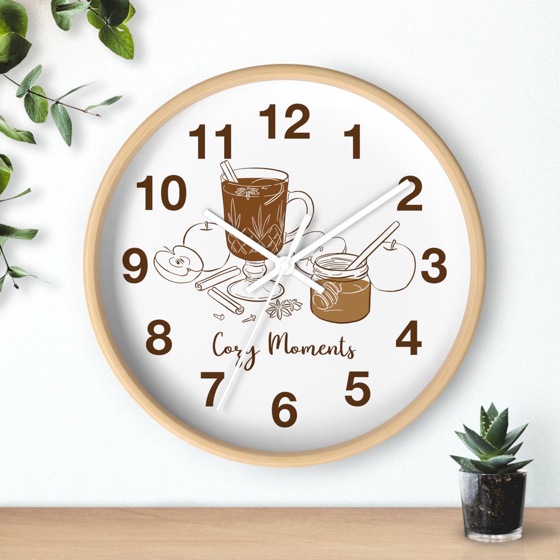 A round wall clock with a light wood frame and a white face, featuring brown numerals and illustrations of a mug, cinnamon sticks, and a jar with the words 'Cozy Moments' on the clock face. The clock is mounted on a white wall above a wooden surface, with decorative green leaves and a small potted succulent nearby.
