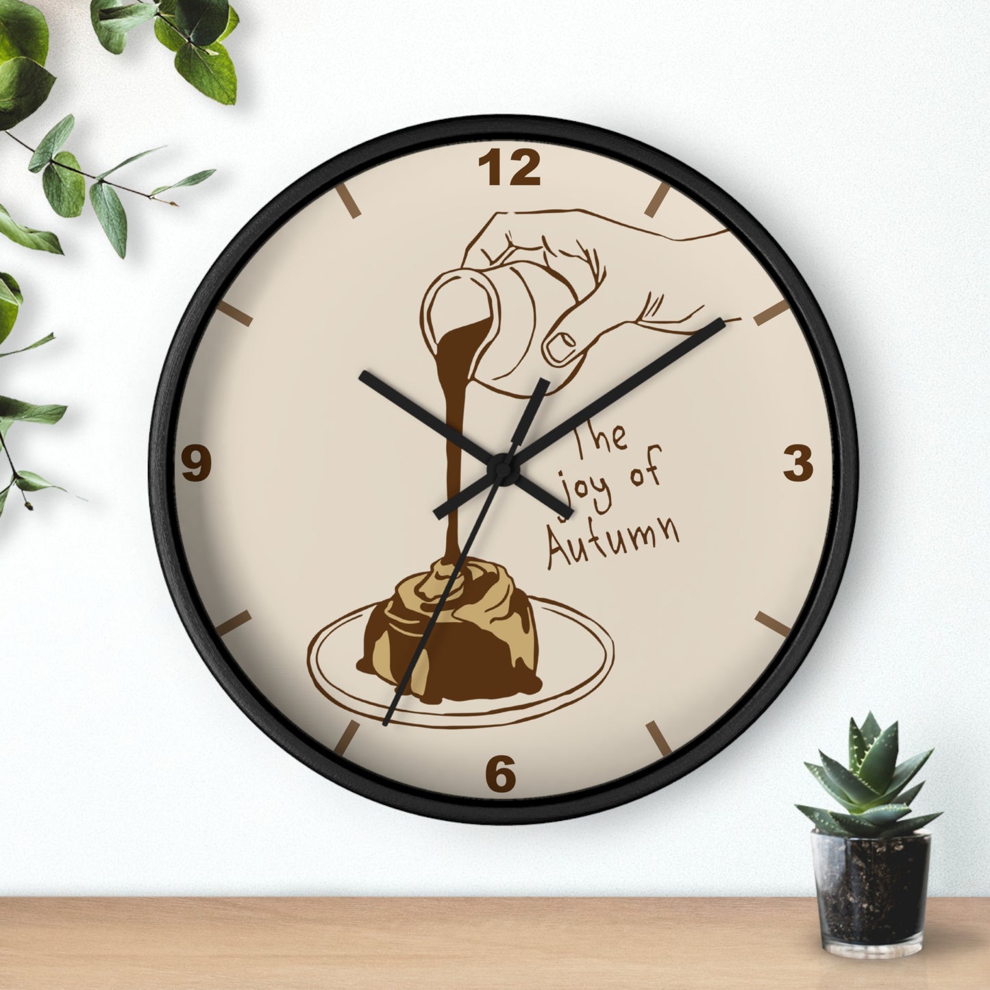 A round wall clock with a black frame and cream face, featuring an illustration of a hand pouring syrup over a dessert with the text 'The joy of Autumn'. The clock is shown hanging on a white wall above a wooden surface, with small green plants in the corners.
