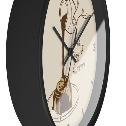 A close-up of a round wall clock with a black frame and a cream-colored face featuring an illustration of chocolate drizzle over a pastry and the text 'The Joy of Autumn'.