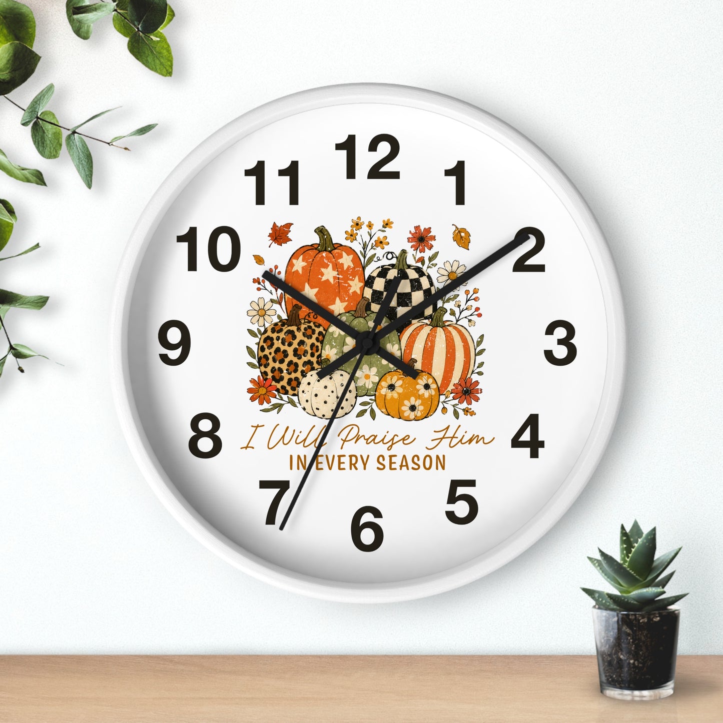 A round white wall clock with a fall harvest design, featuring illustrated pumpkins, autumn leaves, and the text 'I Will Praise Him IN EVERY SEASON'. The clock is shown mounted on a white wall above a light wood surface with decorative green leaves and a small succulent.