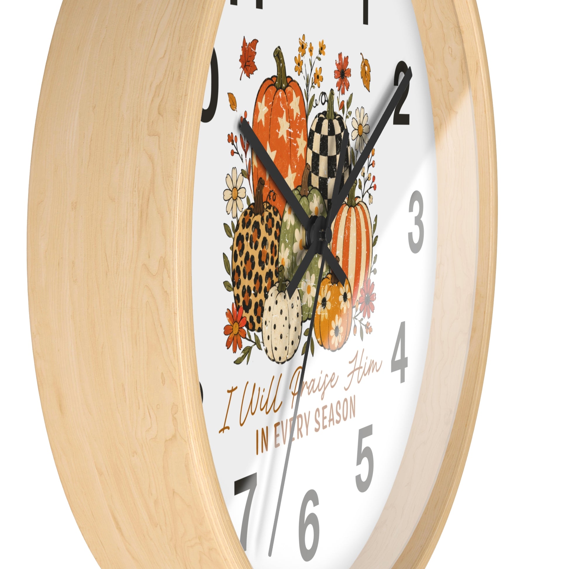 A close-up of a wall clock with a light natural wood frame and a white face featuring a fall harvest illustration of pumpkins, flowers, and the text 'I Will Raise Him IN EVERY SEASON'.