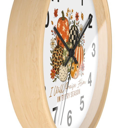 A close-up of a wall clock with a light natural wood frame and a white face featuring a fall harvest illustration of pumpkins, flowers, and the text 'I Will Raise Him IN EVERY SEASON'.