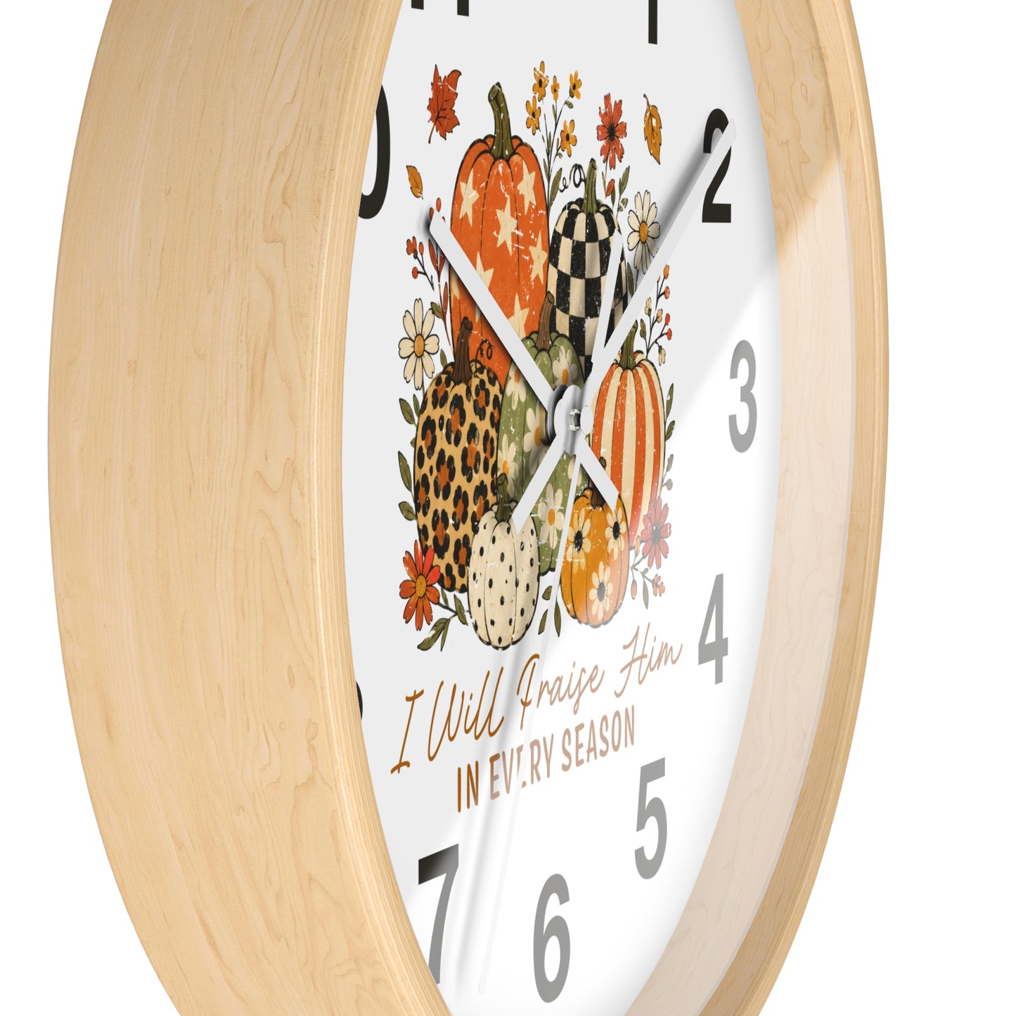 A close-up of a wall clock with a light natural wood frame and a white face featuring a fall harvest illustration with pumpkins, flowers, and the text 'I Will Praise Him IN EVERY SEASON'.