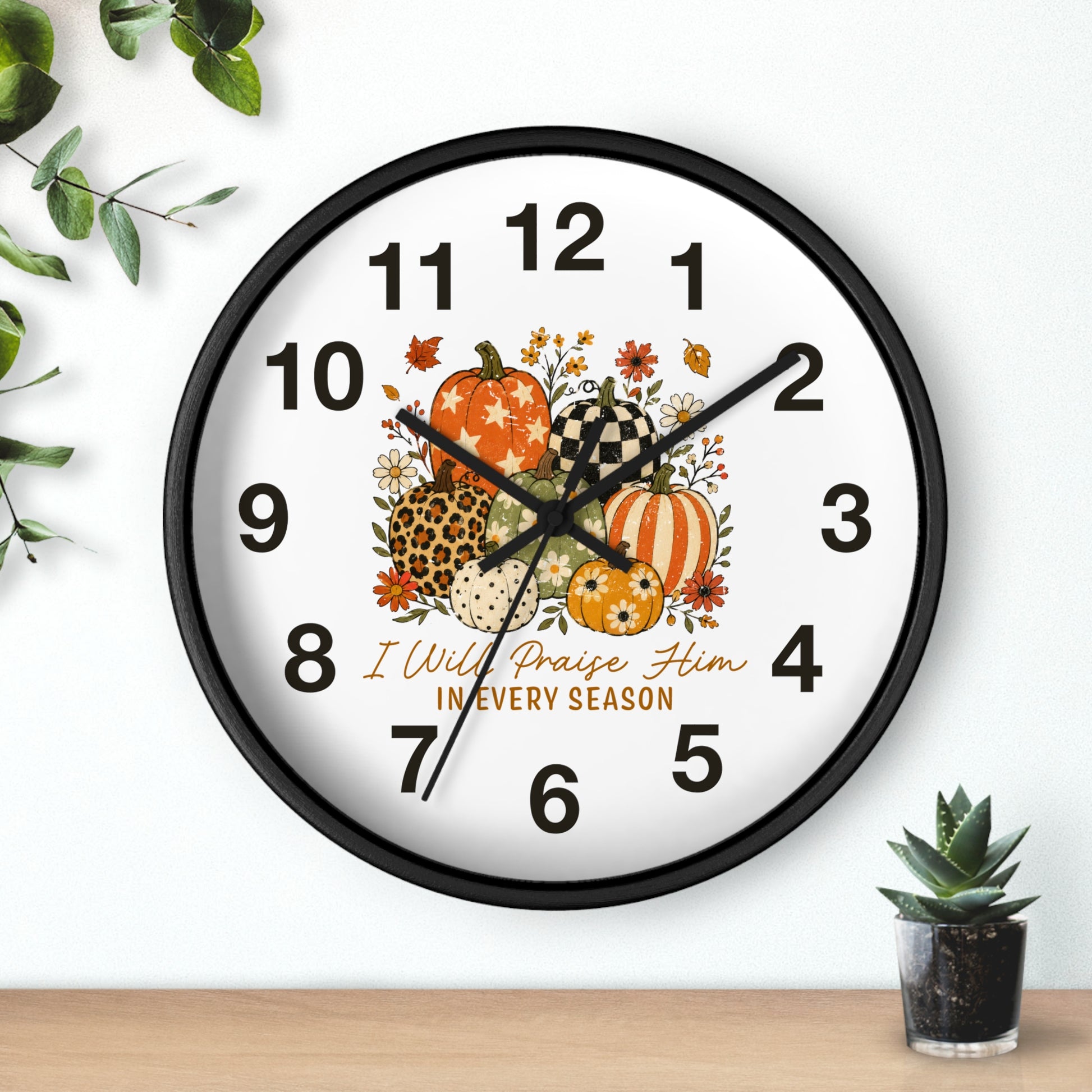 A round wall clock with a black frame and a white face, decorated with autumn pumpkins, flowers, and the text 'I Will Praise Him IN EVERY SEASON.' The clock is shown hanging on a white wall above a wooden surface, with decorative greenery in the corners.