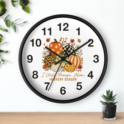 A round wall clock with a black frame and a white face, decorated with autumn pumpkins, flowers, and the text 'I Will Praise Him IN EVERY SEASON.' The clock is shown hanging on a white wall above a wooden surface, with decorative greenery in the corners.
