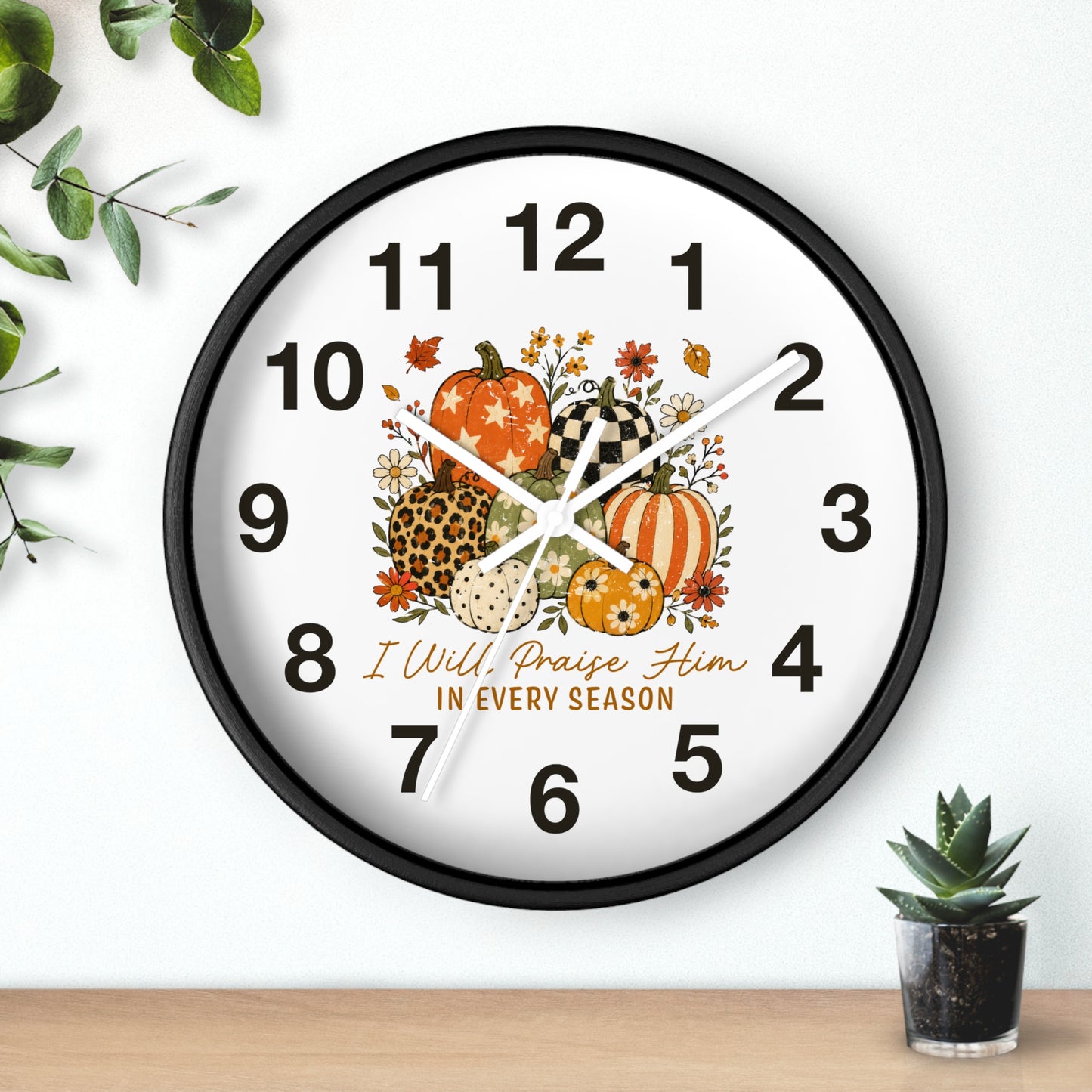 A round wall clock with a black frame and a white face, decorated with autumn pumpkins, flowers, leaves, and the text 'I Will, Praise Him IN EVERY SEASON.' The clock is shown hanging on a white wall above a wooden surface, with a small succulent in a pot at the bottom right and green leaves at the top left.